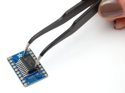A pair of tweezers holding a SOIC chip hovers over the SMT Breakout PCB for SOIC-20 or TSSOP-20.