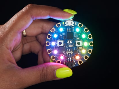 shot of a Black woman's neon-green manicured hand holding up a Circuit Playground Bluefruit glowing rainbow LEDs.