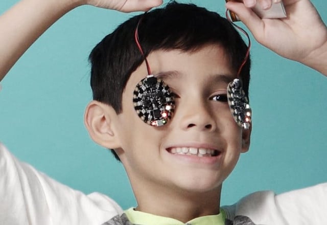 Close shot of two children side by side from just below their shoulders to just above the tops of their heads. Child on left is holding a circuit playground over  each eye with their thumb and index fingers. The back of their hands are facing the camera, covering their cheeks and mouth. Part of this child's face is obscured by the elbow of the child on the right who has their arms in the air and is dangling a circuit playground over each eye. The child's left eye is partially visible. This child is smiling.